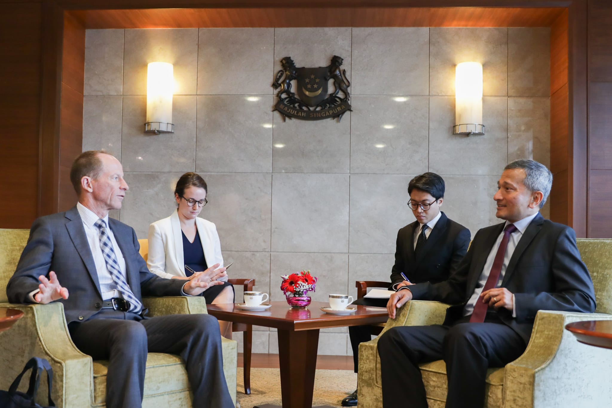 Four people in business attire seated, conversing around a table with cups, beneath Singapore coat of arms.